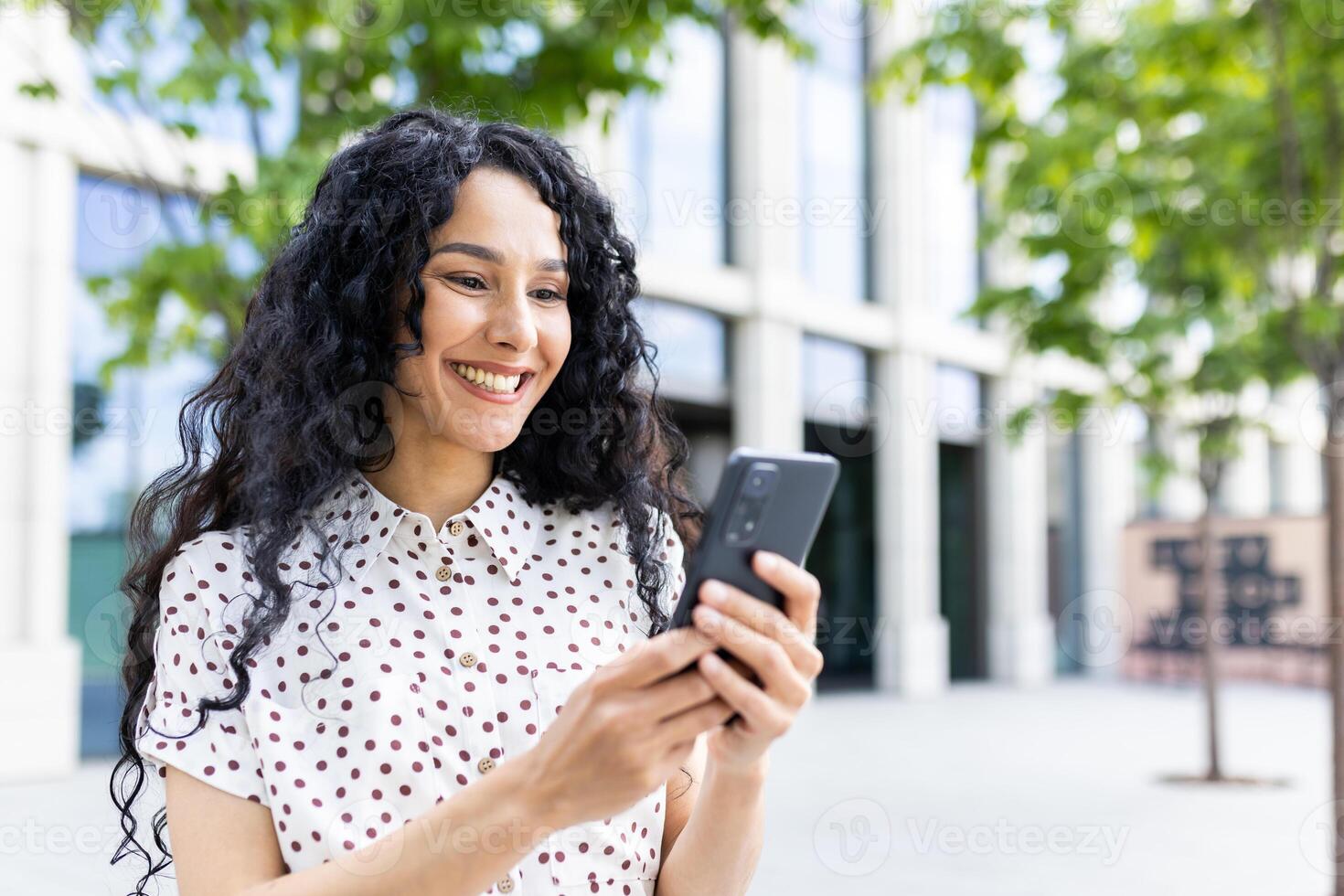 une Jeune magnifique femme des promenades par le ville avec une téléphone dans sa mains, sourit avec contentement, les usages une téléphone intelligent application, parcourt l'Internet pages, les types une texte message, social réseaux. photo