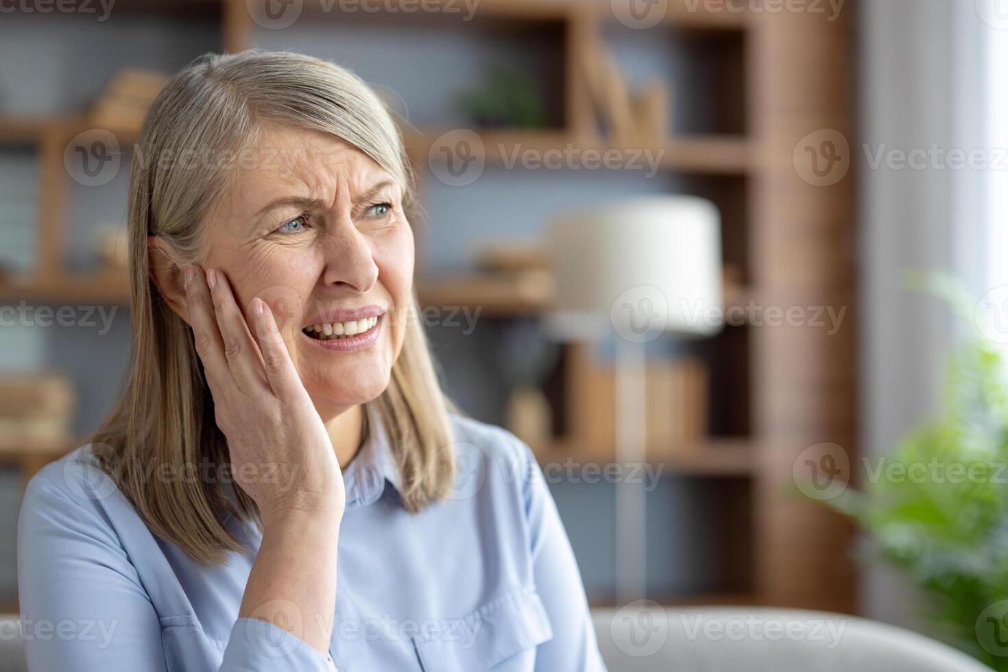 personnes âgées caucasien femme Souffrance de otite, émouvant sa oreille dans douleur, séance dans une moderne vivant chambre. elle regards affligé et inquiet à propos sa mal d'oreille. photo