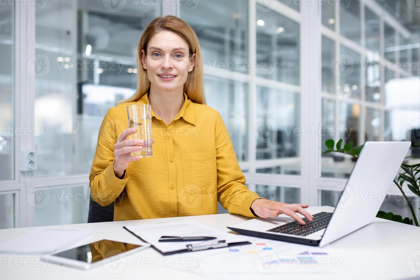 portrait de une Jeune femme dans une Jaune chemise travail dans le Bureau à une portable et en buvant l'eau de une verre, souriant et à la recherche à le caméra. photo