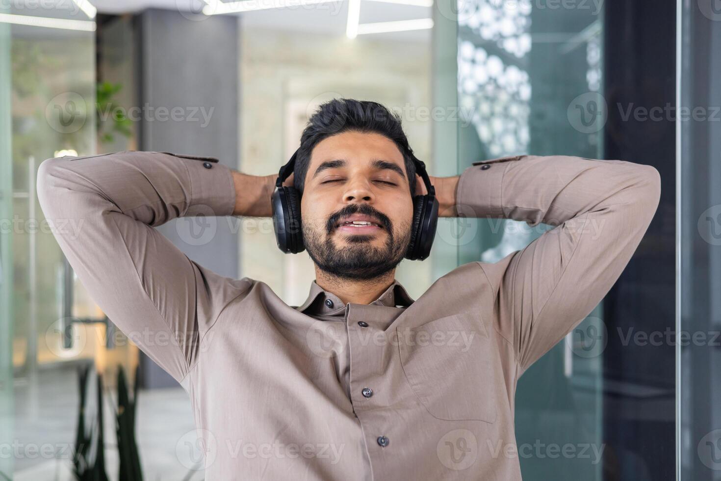 portrait de détendu Indien homme dans écouteurs penché sur retour de chaise avec fermé yeux sur flou Contexte. calme directeur repos dans Bureau avec mains derrière tête et écoute à apaisant playlist. photo