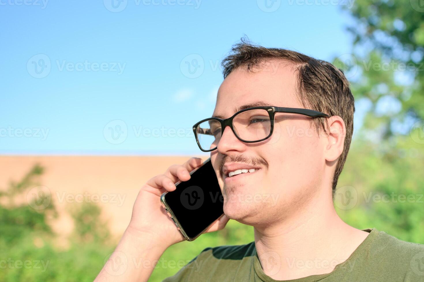 Jeune homme avec lunettes et vert T-shirt parlant sur cellule téléphone à le parc photo