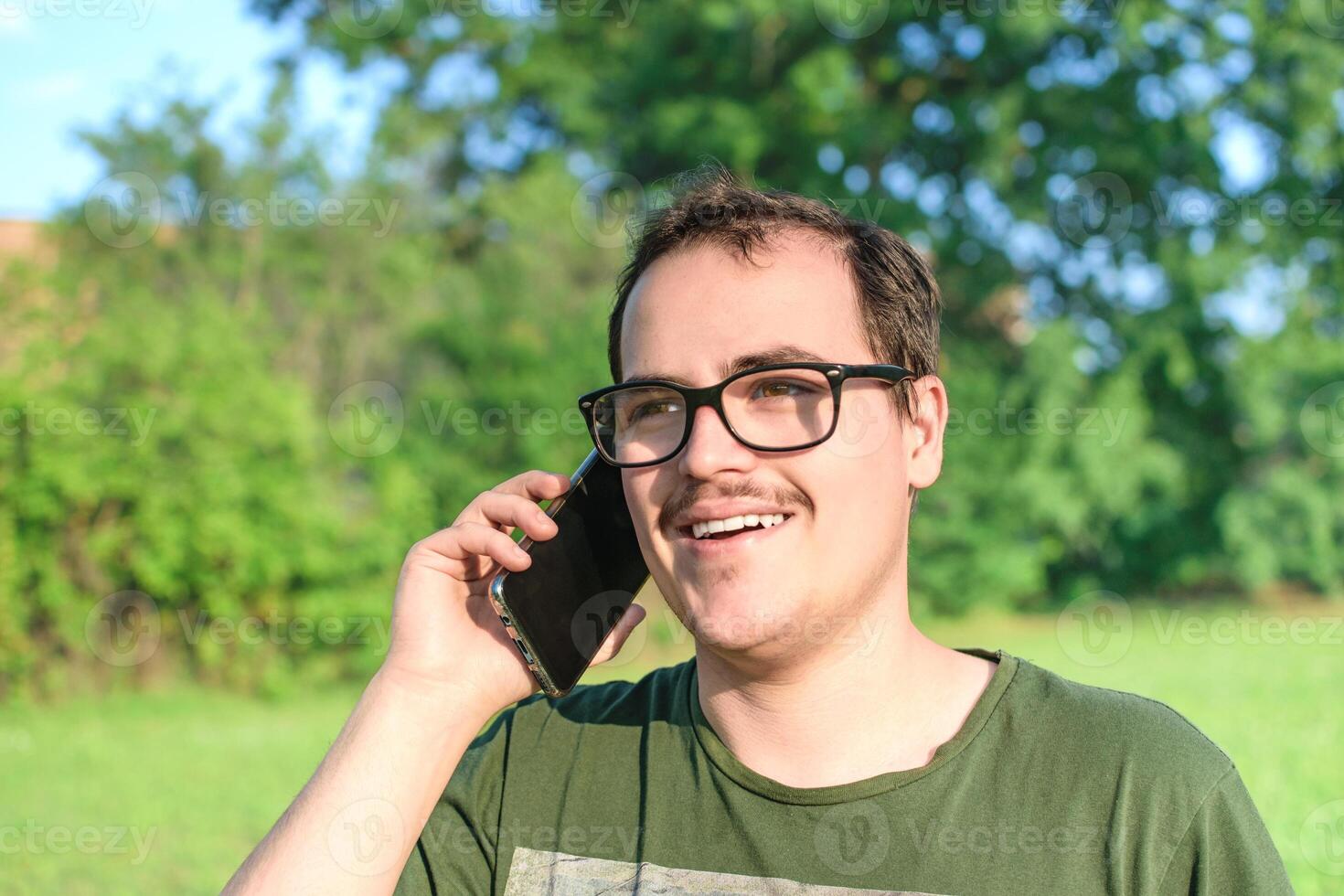 Jeune homme avec lunettes et vert T-shirt parlant sur cellule téléphone à le parc photo