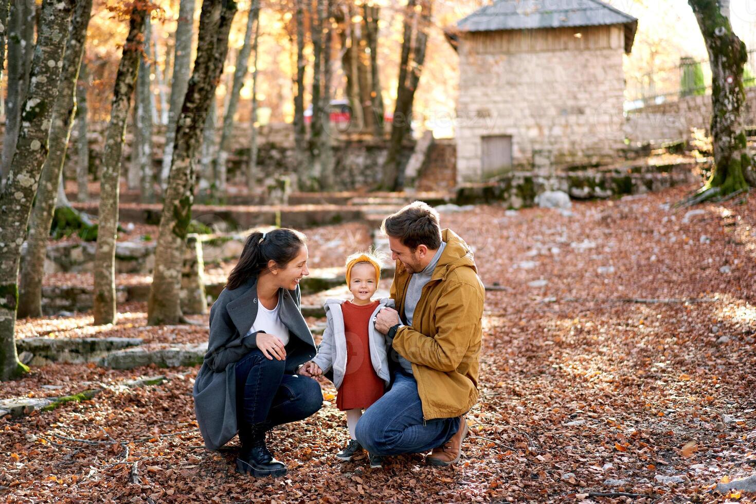 souriant papa et maman sont squat près une peu fille parmi le déchue feuilles dans le forêt photo