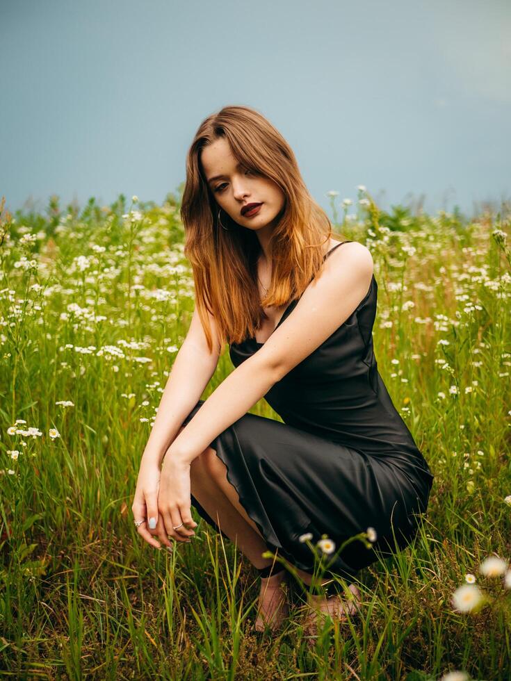 magnifique Jeune fille dans une noir soir robe pose accroupi dans de face de une Marguerite champ sur une nuageux été journée. portrait de une femelle modèle en plein air. pluvieux temps. gris des nuages. photo