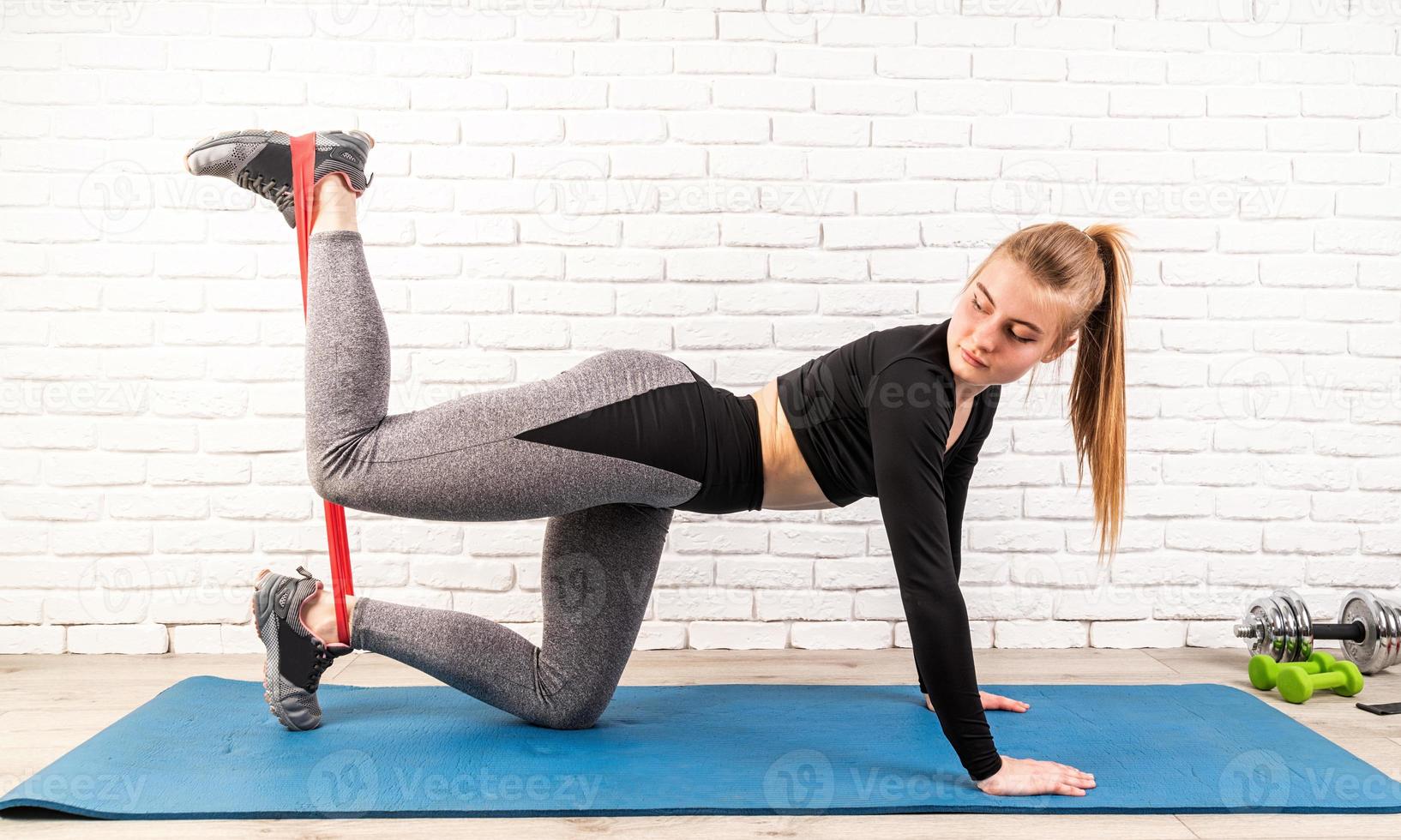 jeune sportive faisant les exercices à quatre pattes, cambrant le dos et redressant la jambe à la maison à l'aide d'une bande de résistance en caoutchouc photo