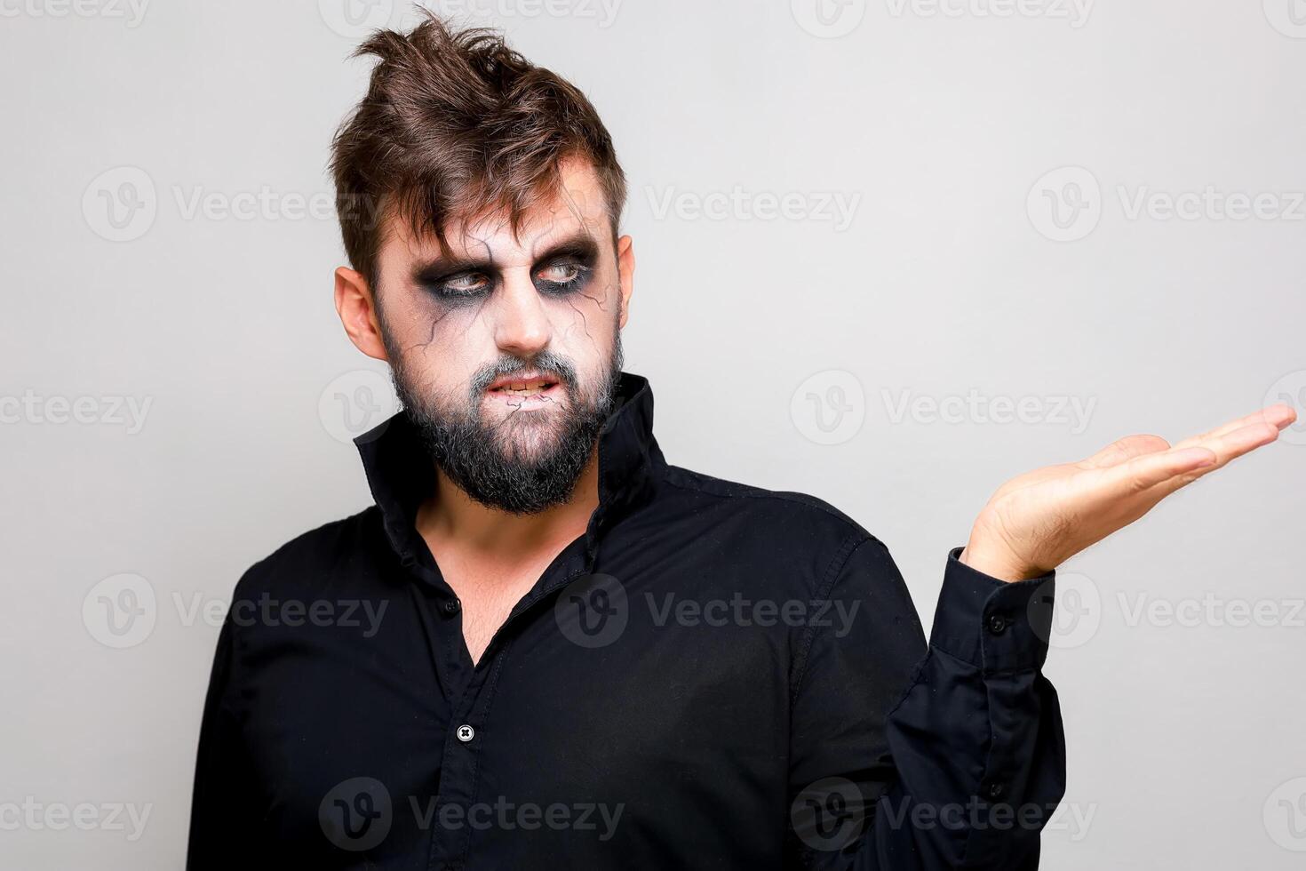 une homme avec une barbe et Mort-vivant maquillage sur Halloween détient le sien mains dans de face de lui photo