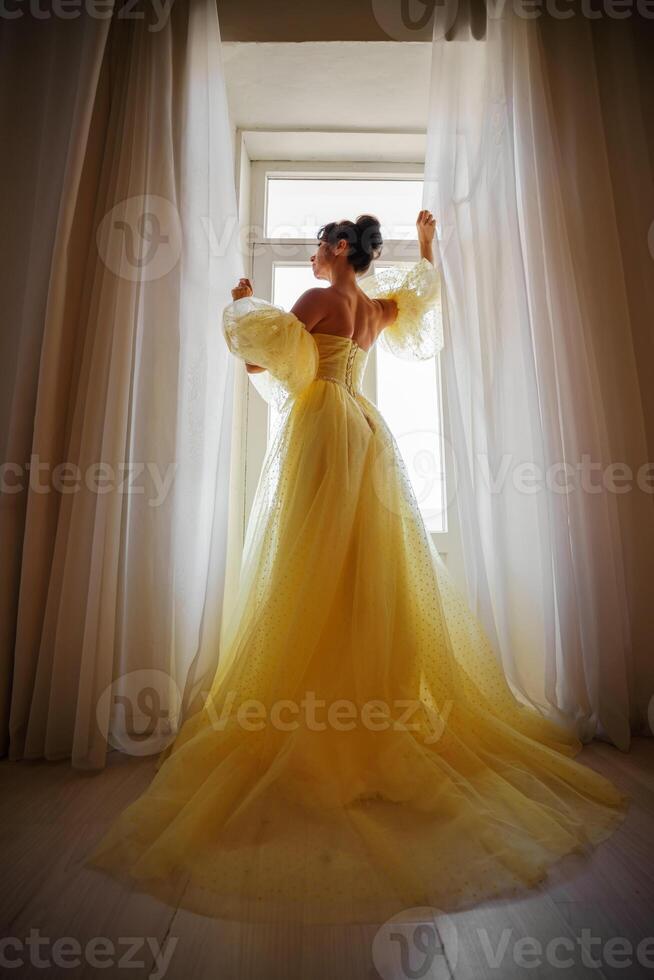 une femme silhouette dans une d'or luxueux robe contre le Contexte de une fenêtre détient une rideau avec sa mains. élégant Dame dans une Jaune longue soie robe avec nu dos, retour voir. photo