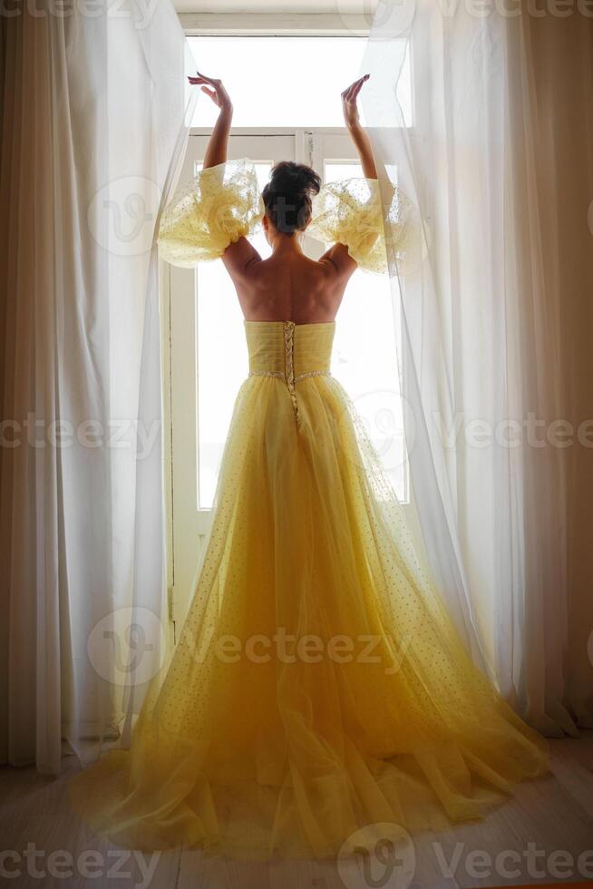 une femme silhouette dans une d'or luxueux robe contre le Contexte de une fenêtre détient une rideau avec sa mains. élégant Dame dans une Jaune longue soie robe avec nu dos, retour voir. photo