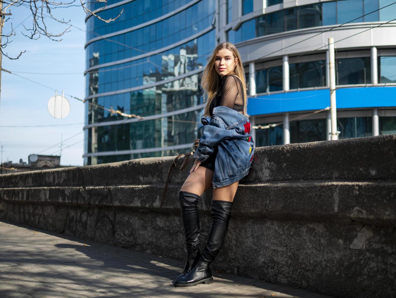 fille en bottes de cuir et une veste en jean se dresse dans le contexte d'un centre d'affaires moderne photo