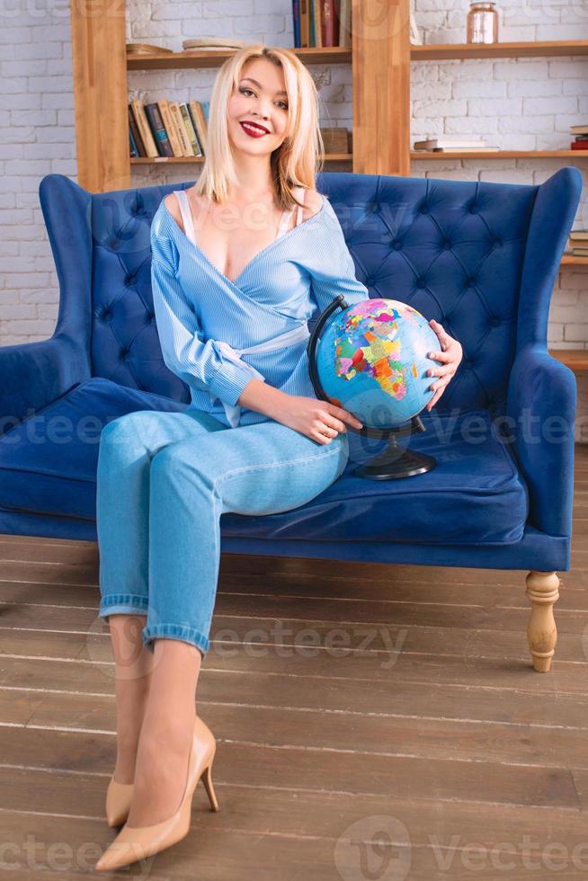Portrait d'une belle femme slave blonde gaie et séduisante en jean bleu, chemisier bleu assis sur le canapé bleu tenant un globe terrestre et rêvant de voyager photo