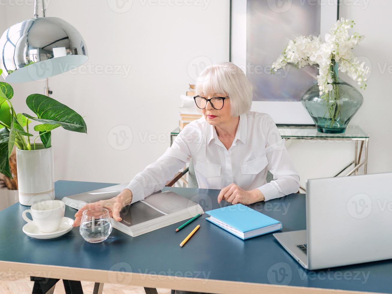 femme senior aux beaux cheveux gris en blouse blanche buvant de l'eau pendant le travail au bureau. travail, personnes âgées, bilan hydrique, trouver une solution, concept d'expérience photo