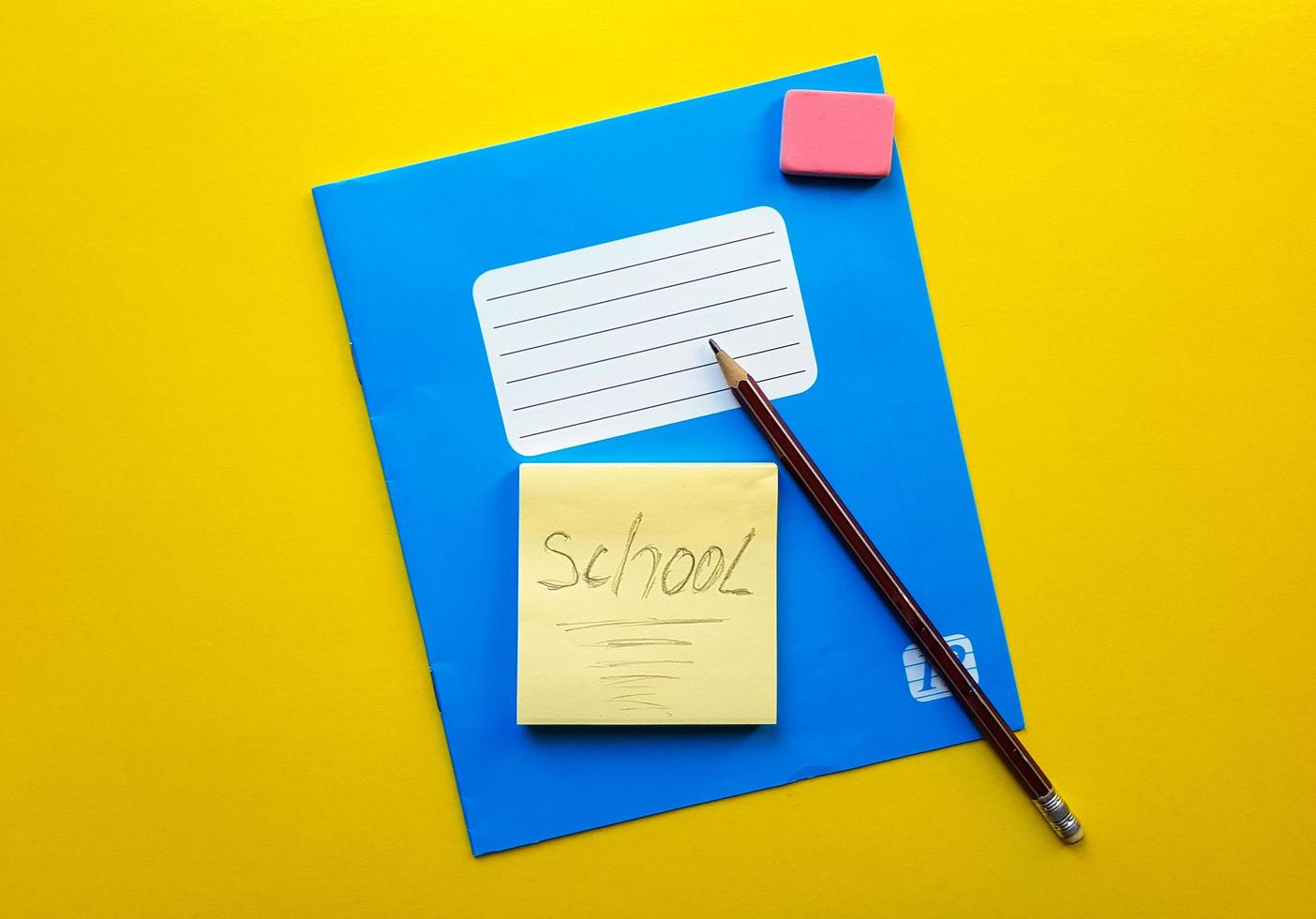 cahier bleu et bout de copie de manuel pour les matières scolaires, gomme, crayon, trombone sur fond jaune. le mot école est écrit au crayon. mise à plat, espace de copie, vue de dessus, place pour le texte. photo