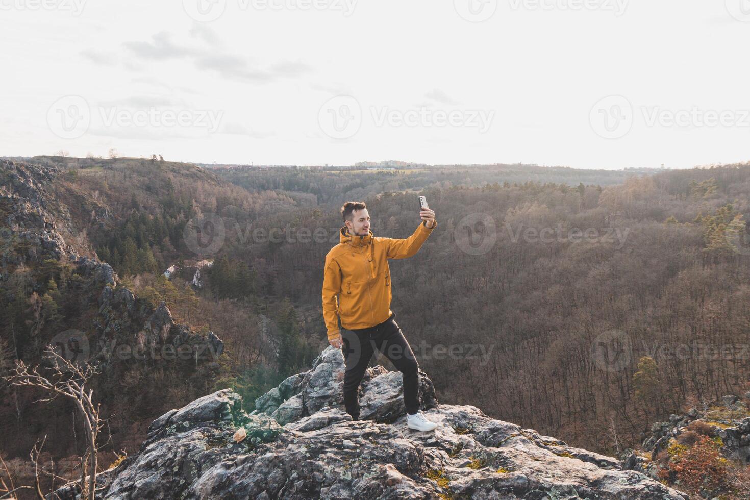 voyageur dans une Jaune veste permanent sur le bord de une falaise prise une selfie sur le sien mobile téléphone à le coucher du soleil. divoka sarka vallée, Prague photo