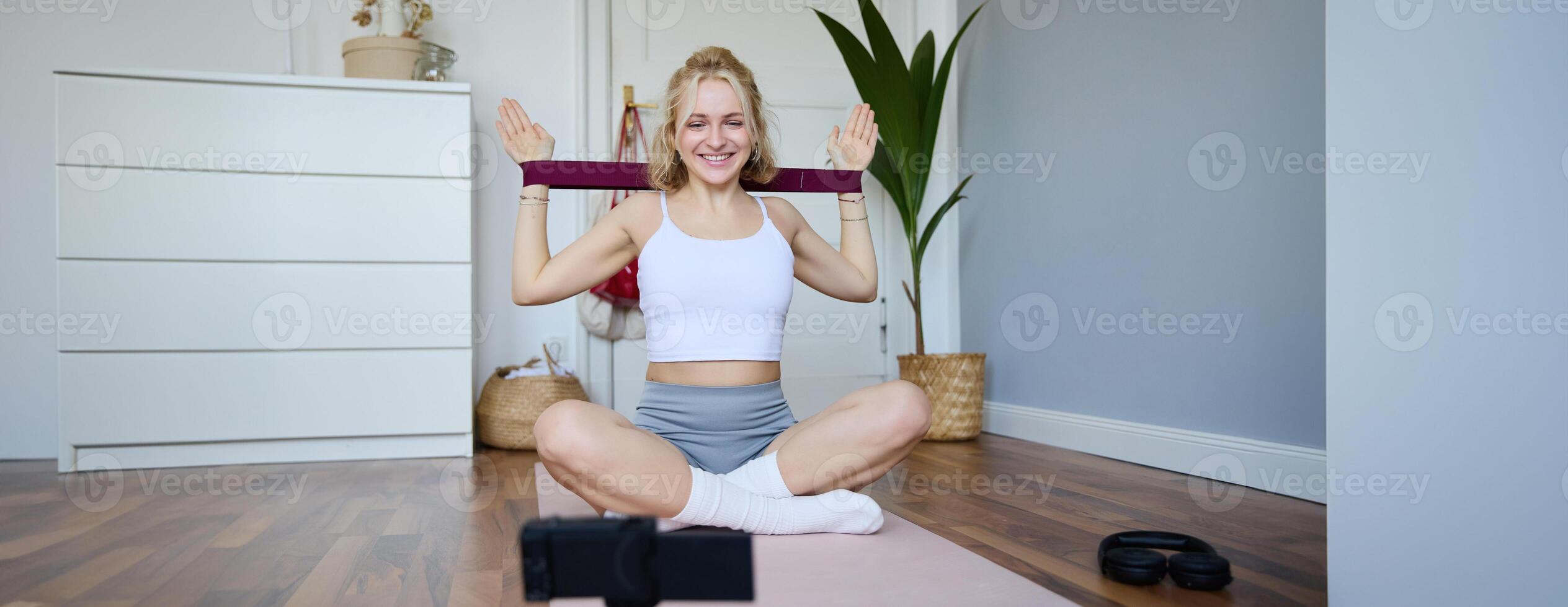 portrait de Jeune souriant femme, aptitude instructeur enregistrement à propos entraînement, montrant Comment à exercice à Accueil et utilisation caoutchouc la résistance groupe, séance sur yoga tapis photo