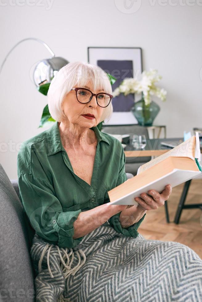 joyeuse femme âgée assise sur le canapé en lisant un livre à la maison. éducation, maturité, concept de loisirs photo