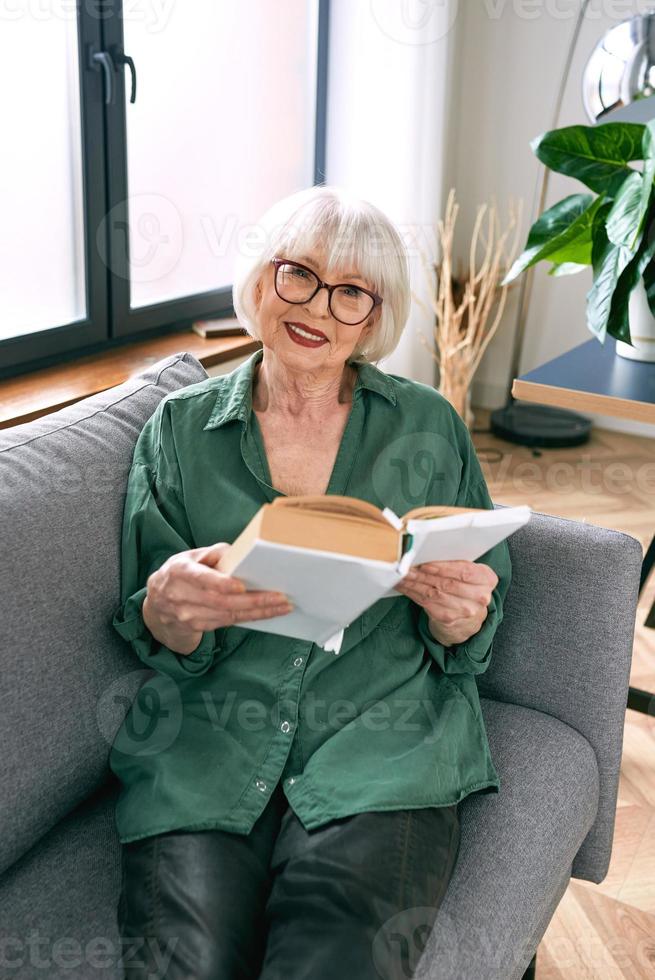joyeuse femme âgée assise sur le canapé en lisant un livre à la maison. éducation, maturité, concept de loisirs photo