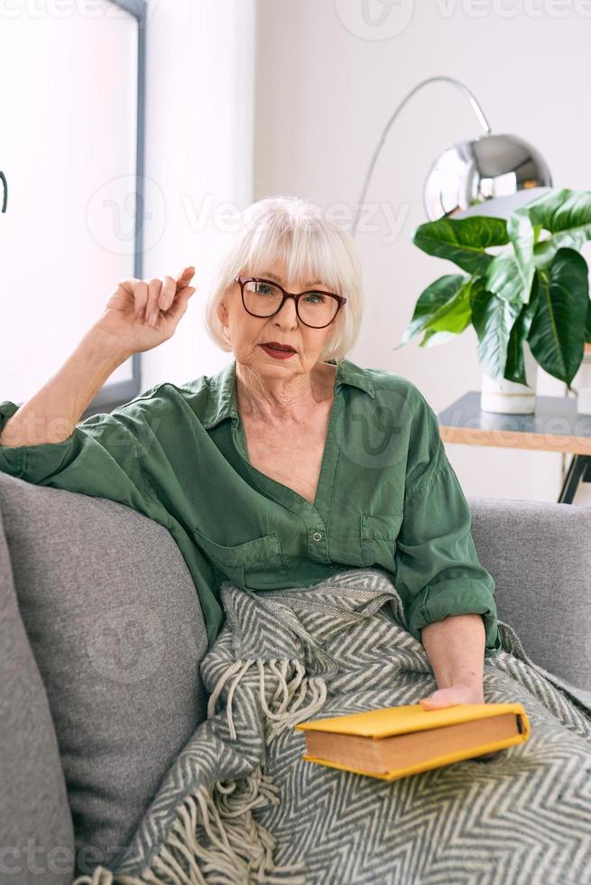 joyeuse femme âgée assise sur le canapé en lisant un livre à la maison. éducation, maturité, concept de loisirs photo