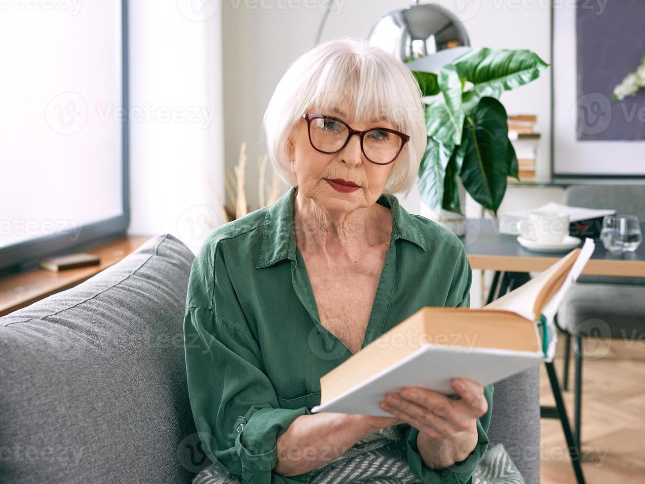 joyeuse femme âgée assise sur le canapé en lisant un livre à la maison. éducation, maturité, concept de loisirs photo