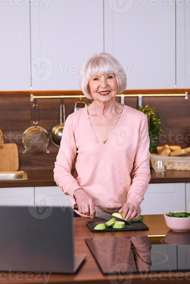 femme joyeuse senior cuisine dans une cuisine moderne par ordinateur portable. nourriture, éducation, concept de mode de vie photo