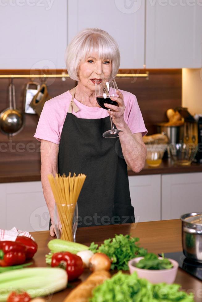 une femme âgée et joyeuse boit du vin rouge pendant la cuisson dans une cuisine moderne. nourriture, éducation, concept de mode de vie photo