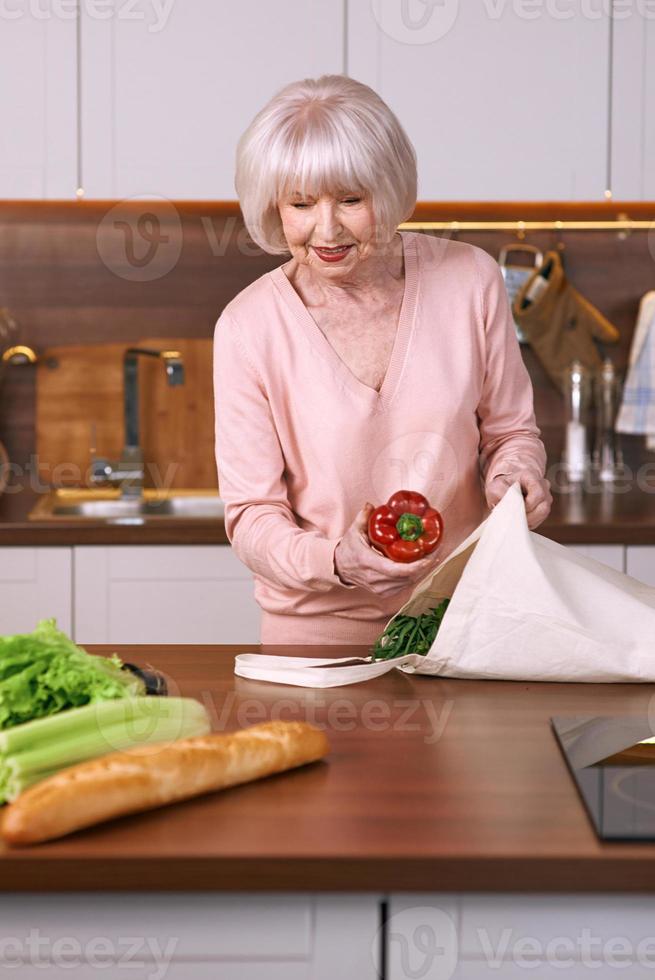 une femme joyeuse et âgée remballe l'épicerie dans une cuisine moderne. nourriture, compétences, concept de mode de vie photo