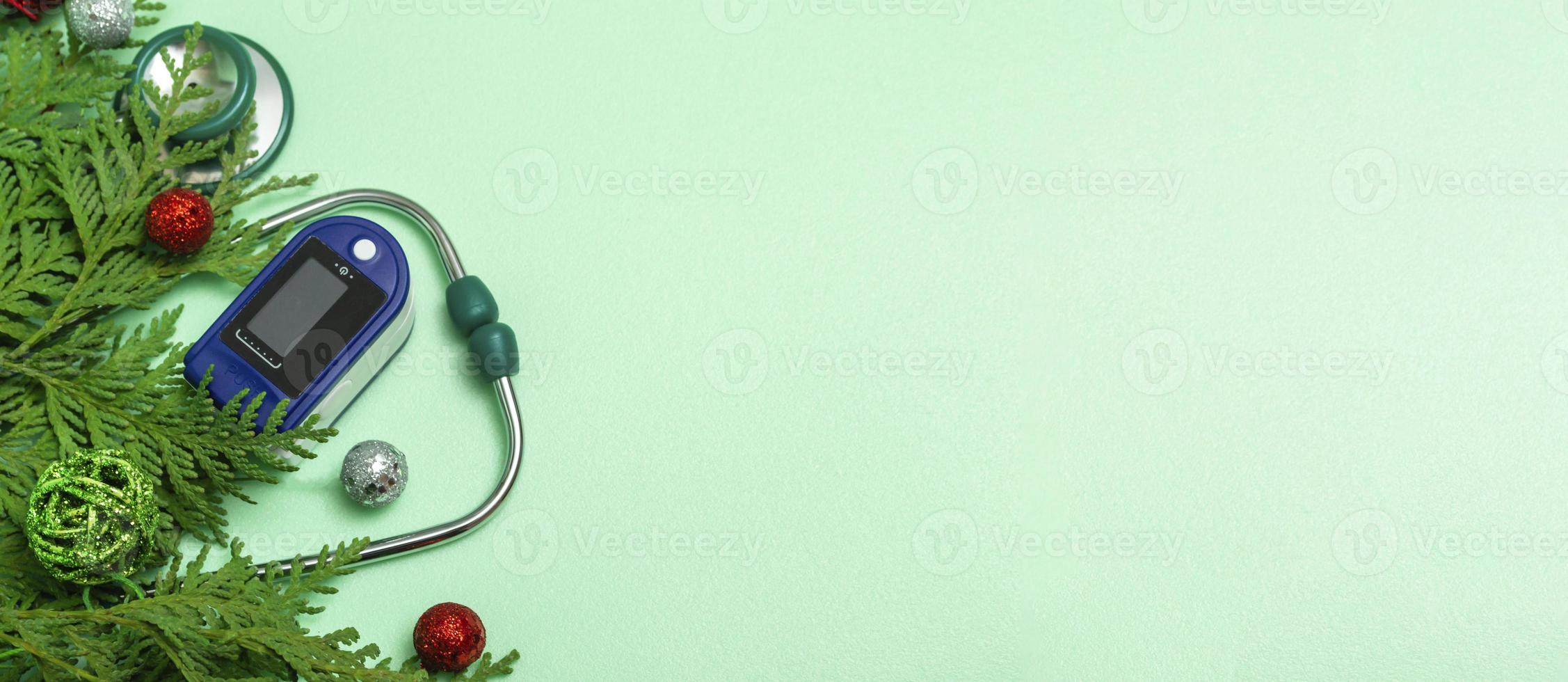 concept médical célébrant Noël dans les soins de santé. vue de dessus d'un gros plan à plat d'un stéthoscope, oxymètre de pouls avec des branches de sapin sur fond vert. espace de copie photo