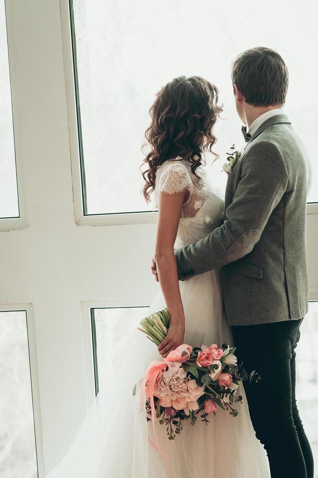 mariée et le marié avec bouquet de mariage debout près de la grande fenêtre, vue arrière photo