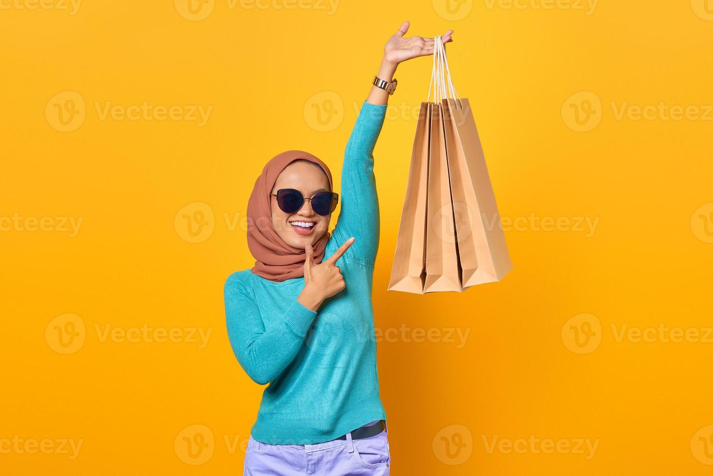 Souriante jeune femme asiatique pointant du doigt les sacs à provisions sur fond jaune photo