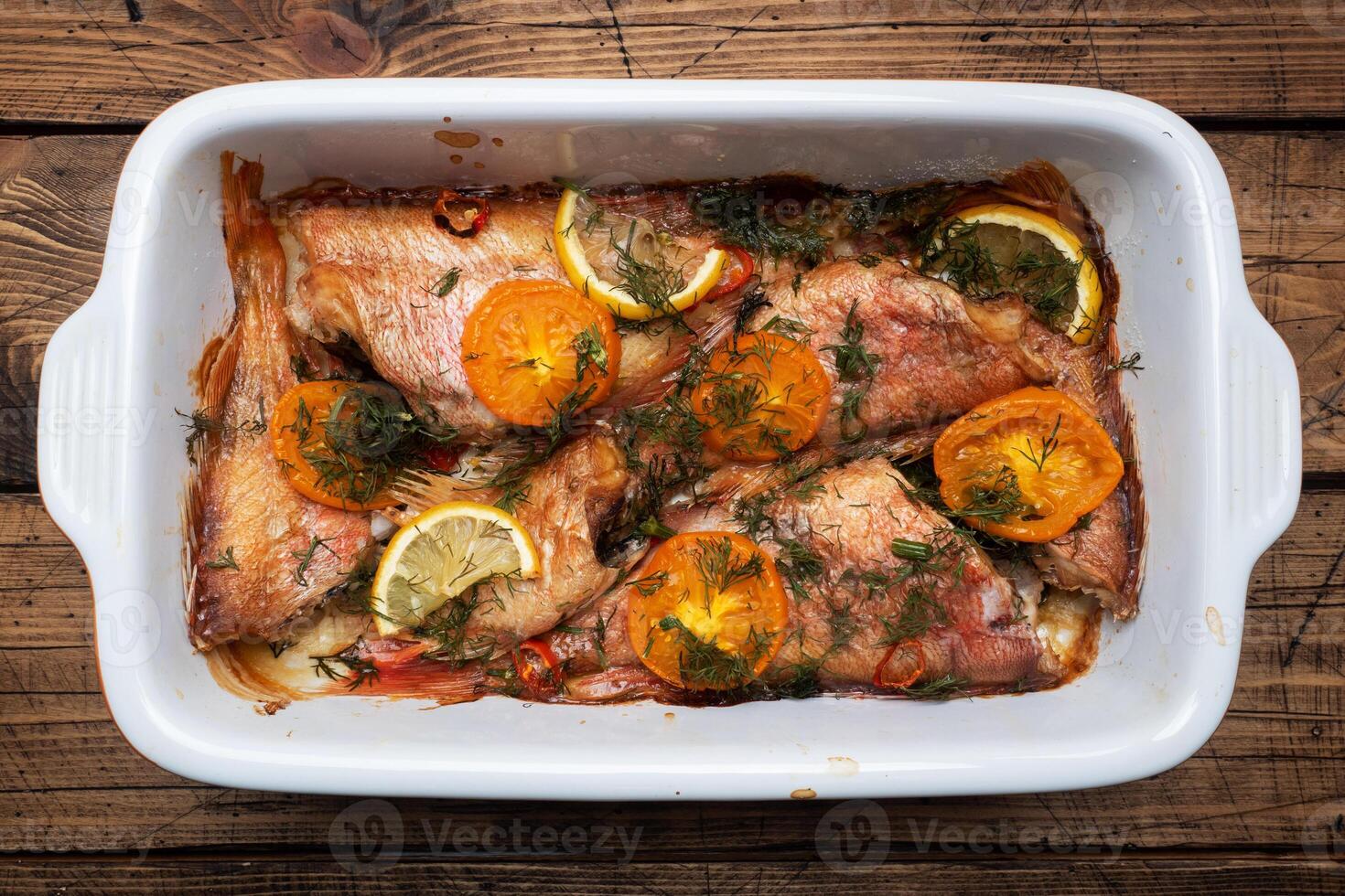 cuit mer basse avec tomate poivre citron et herbes dans une céramique cuisson feuille sur une en bois Contexte. poisson cuit dans une Accueil four sur le tableau. photo