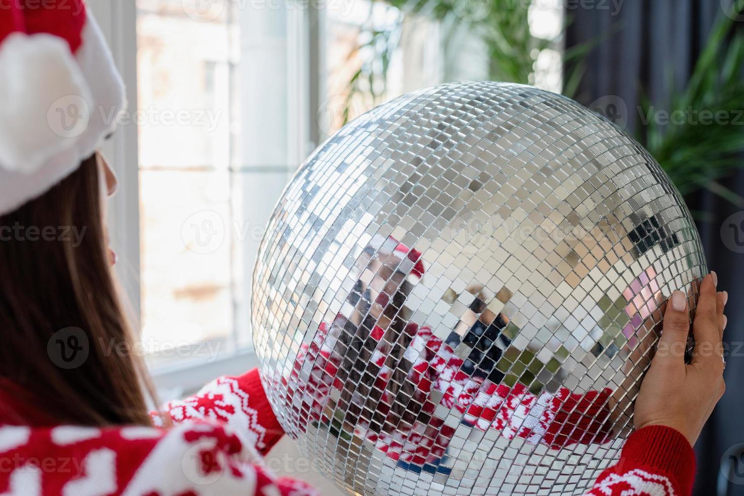 Jeune femme en bonnet de noel tenant une boule disco miroir debout près de l'arbre de Noël photo