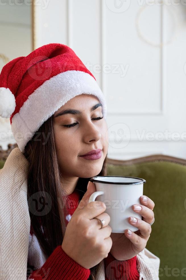 Jeune femme assise sur un canapé seule dans un salon décoré pour Noël ayant une tasse de boisson chaude photo