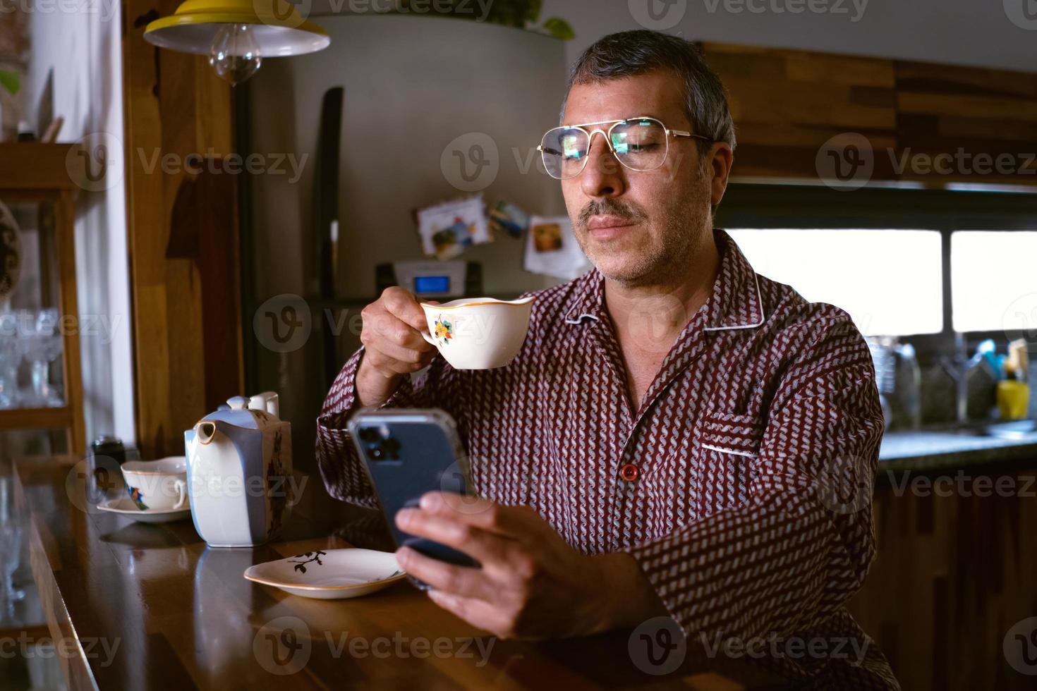 un homme à la maison boit un café en vérifiant son téléphone portable photo