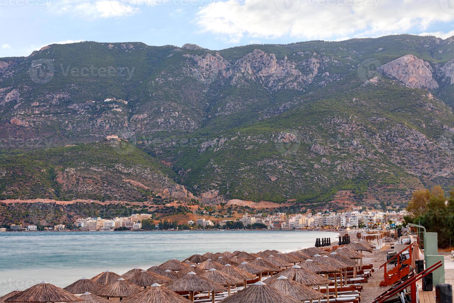 vue sur les pics de chaume des parasols et des chaises longues en bois avec matelas sur le front de mer désert de loutraki en grèce sur fond de montagnes tôt le matin. photo