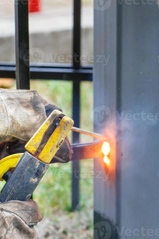 les mains d'un soudeur dans des gants de cuir rugueux avec un porte-électrode soudent une clôture métallique. photo