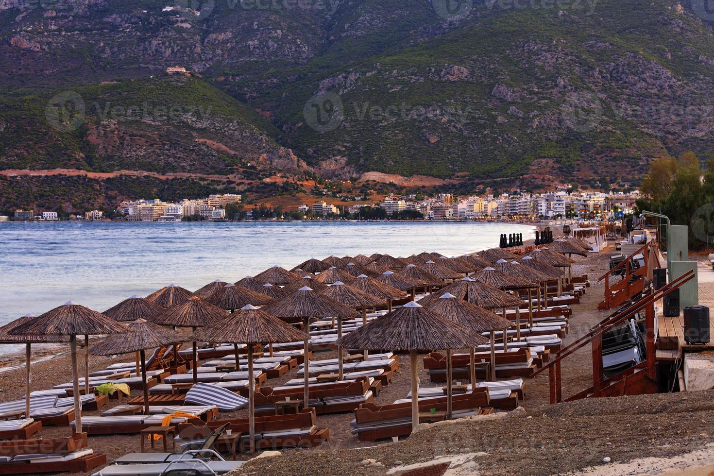 des pics de chaume de parasols et de transats en bois avec matelas sur une promenade déserte parmi les montagnes et les monastères au loin dans les rayons du soleil couchant de la ville. photo