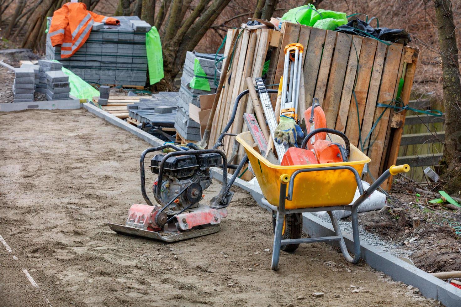 une brouette de construction est remplie de divers outils sur un chantier de construction à proximité d'une plaque vibrante à essence et de piles de dalles de pavage. photo