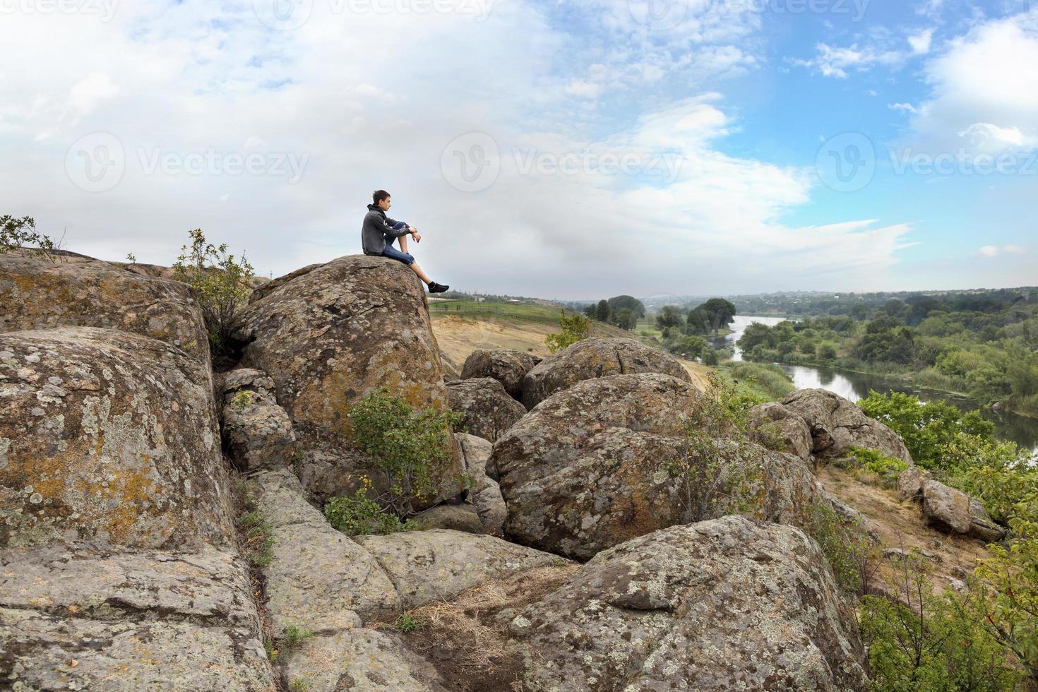 l'adolescent est assis au sommet d'un gros rocher de pierre sur la rive de la rivière bug sud et regarde la rivière en contrebas photo