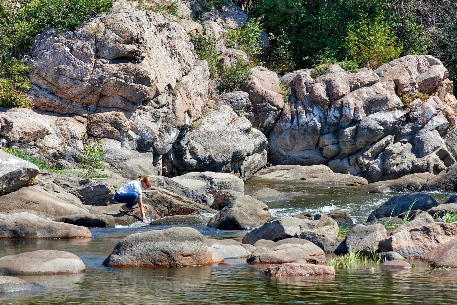 une jeune femme plonge ses mains dans les jets d'eau mousseux sur des rochers de pierre au bord d'une rivière forestière par une journée d'été ensoleillée. photo