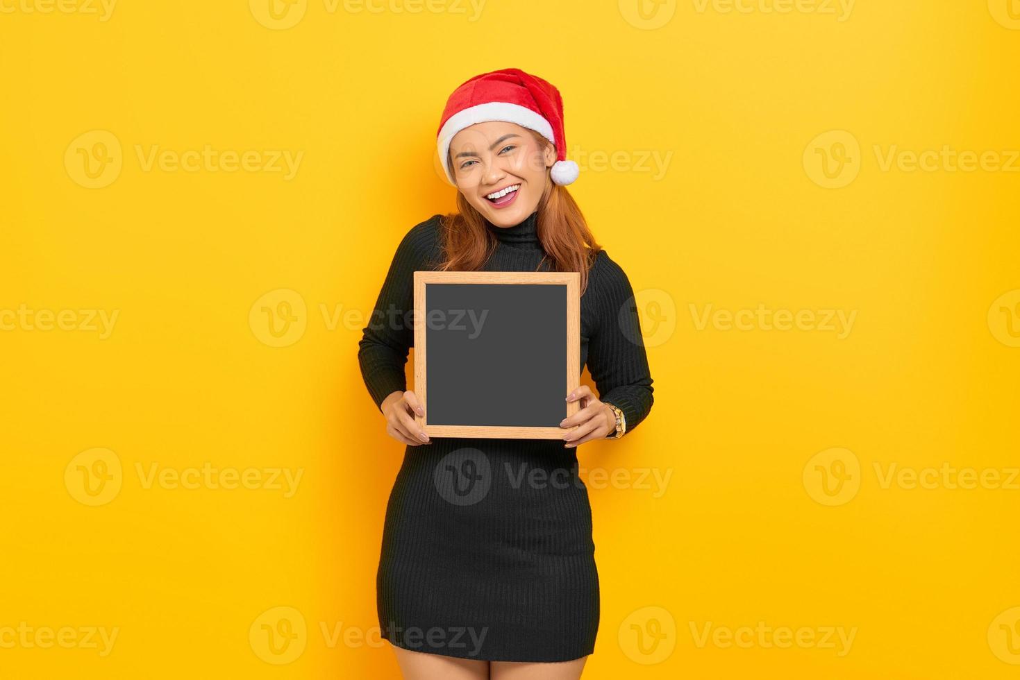 Portrait of smiling young asian woman in santa claus hat holding tableau isolé sur fond jaune photo