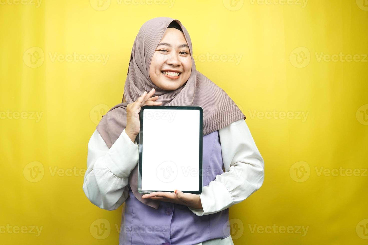 belle jeune femme musulmane asiatique souriante, excitée et joyeuse tenant une tablette avec un écran blanc ou vierge, faisant la promotion de l'application, faisant la promotion du produit, présentant quelque chose, isolée sur fond jaune photo