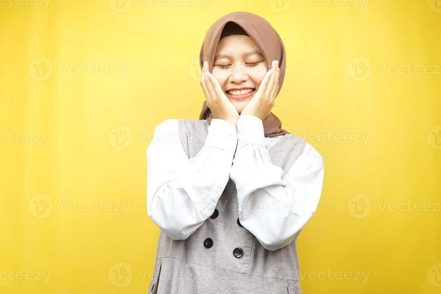 belle jeune femme musulmane asiatique souriante heureuse, mignonne, se sentant à l'aise, se sentant soignée, se sentant bien, avec les mains tenant les joues isolées sur fond jaune photo