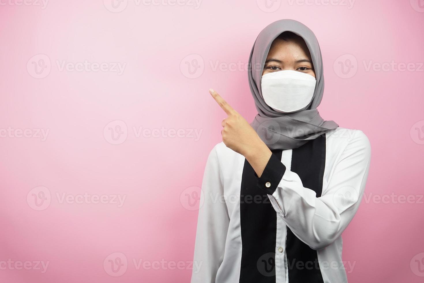 femme musulmane portant un masque blanc, la main pointant vers un espace vide présentant quelque chose, isolé sur fond rose photo