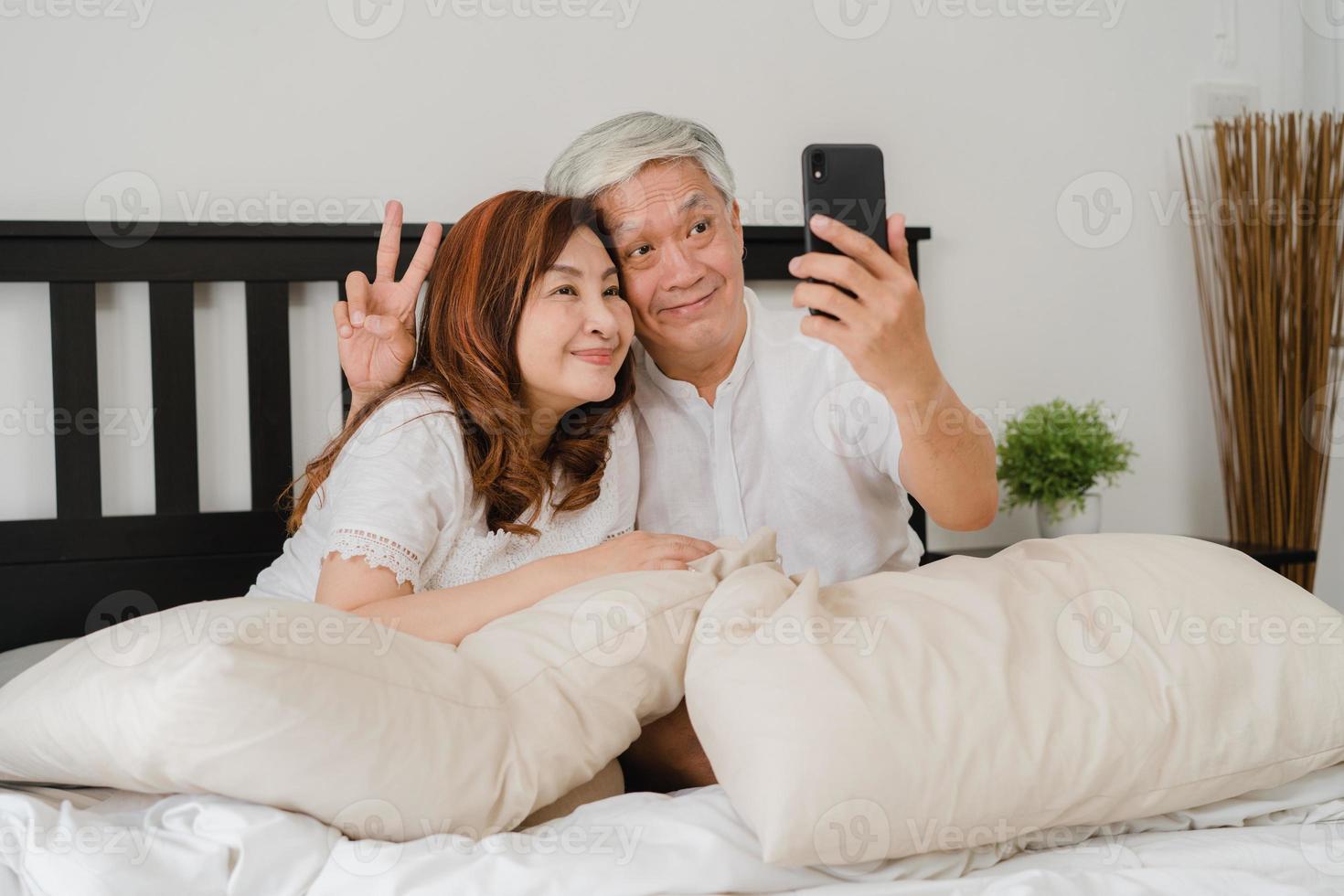 selfie de couple de personnes âgées asiatiques à la maison. grands-parents chinois seniors asiatiques, mari et femme heureux d'utiliser un téléphone portable selfie après s'être réveillé allongé sur le lit dans la chambre à la maison le matin concept. photo