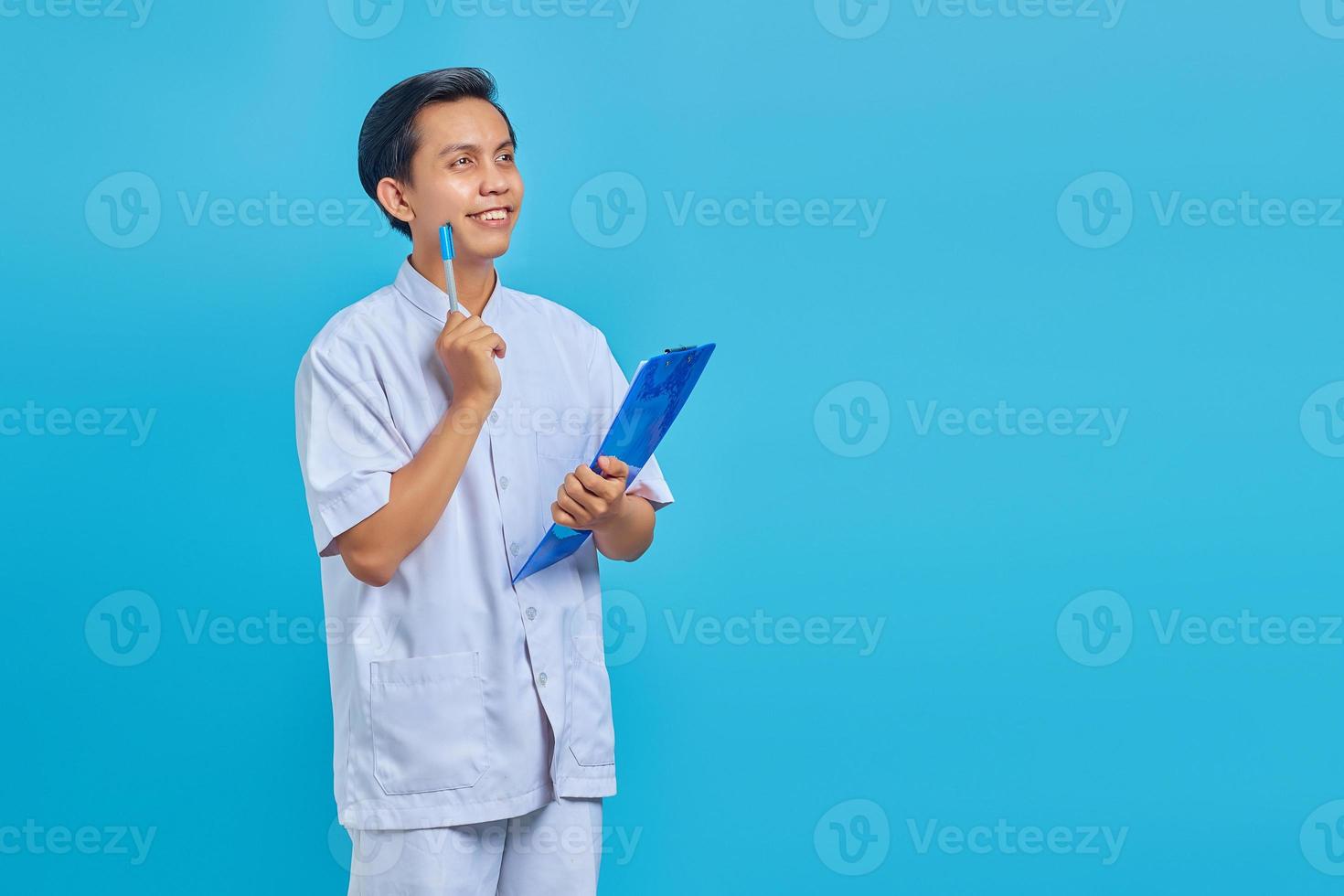 beau jeune infirmier se concentrant en pensant à des idées et souriant en regardant un espace vide sur fond bleu photo