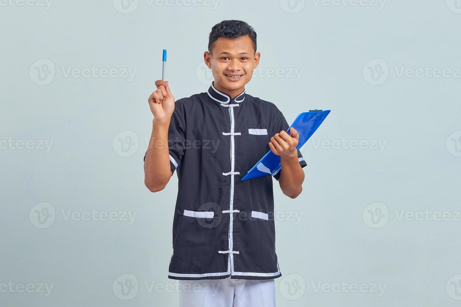 Portrait d'un jeune homme asiatique souriant portant un uniforme de karaté tenant un presse-papiers et pointant vers le haut avec un stylo photo