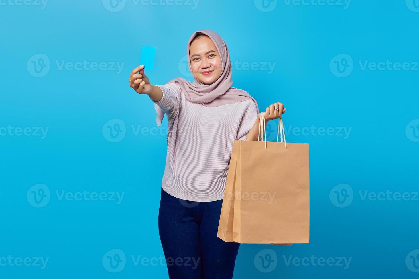 Portrait d'une femme asiatique joyeuse tenant un sac à provisions et montrant une carte de crédit sur fond bleu photo