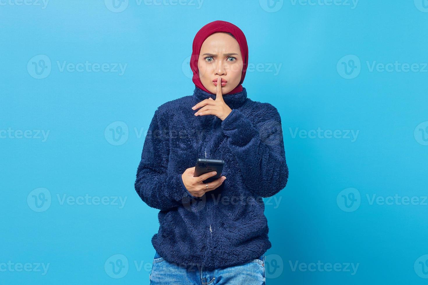 belle jeune femme asiatique tenant un téléphone portable et fait un geste de silence sur fond bleu photo