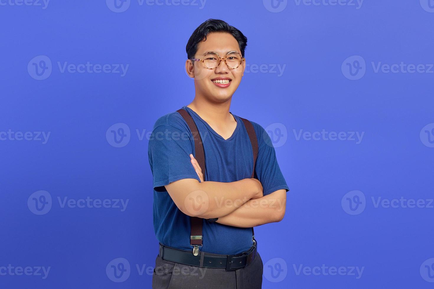 portrait d'un jeune homme asiatique souriant croisant les bras et portant des lunettes sur fond violet photo