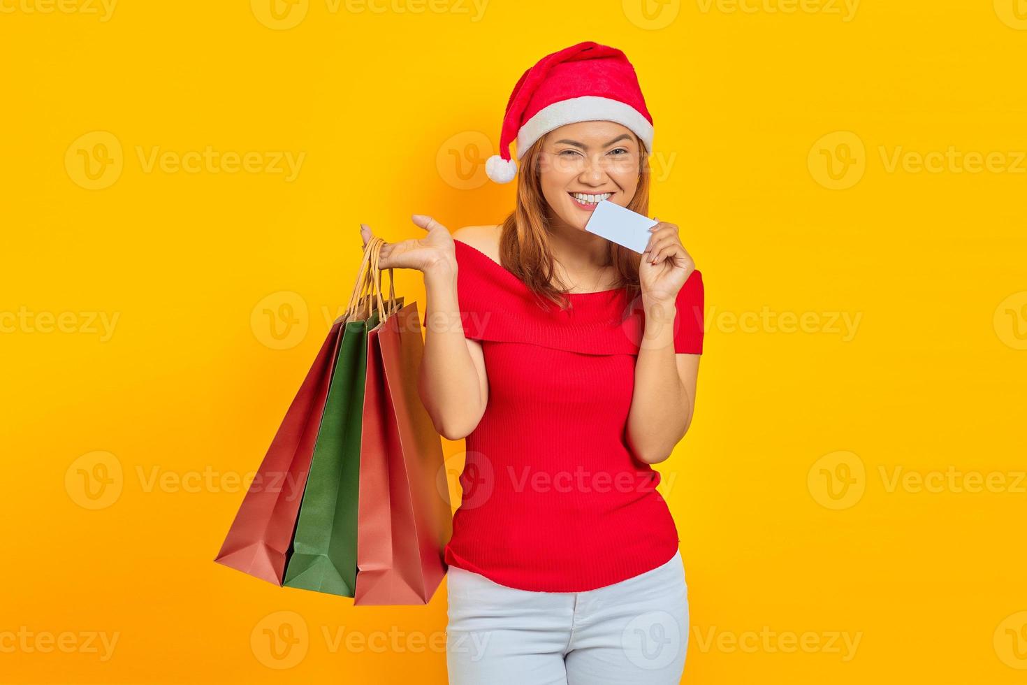 Souriante jeune femme asiatique en chapeau de père Noël tenant un sac à provisions et mordre la carte de crédit sur fond jaune photo