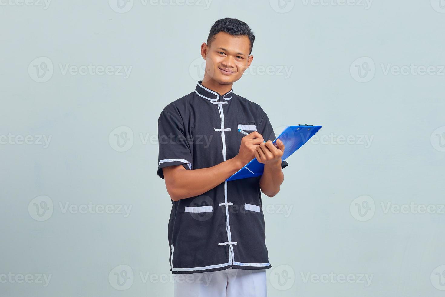 Portrait d'un jeune homme asiatique souriant portant un uniforme de karaté prenant des notes sur le presse-papiers sur fond gris photo